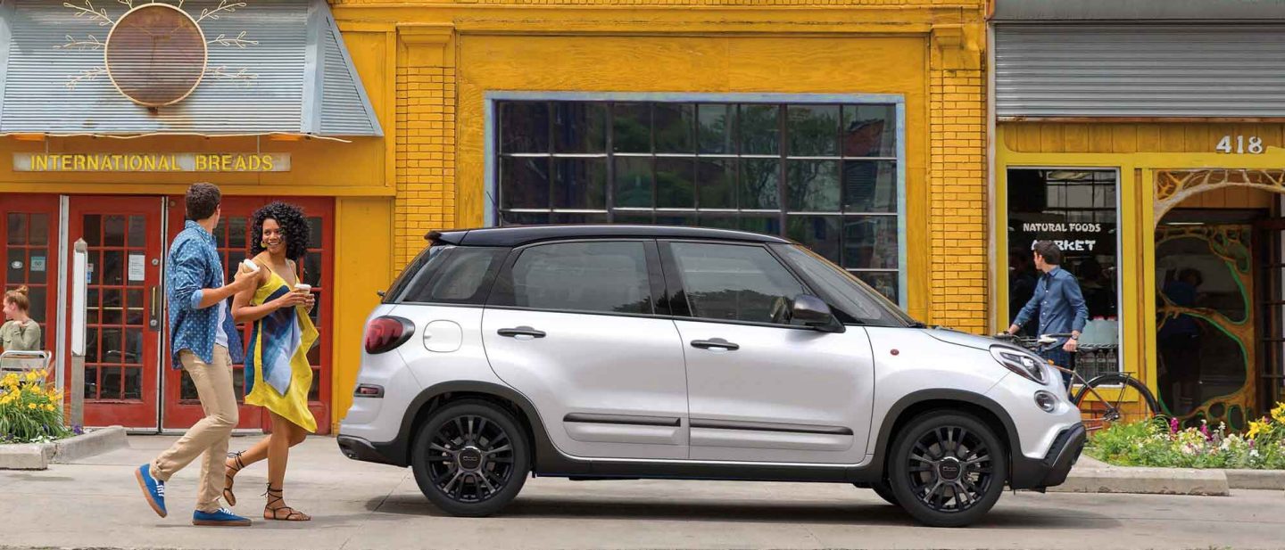 A man and woman in conversation approach a 2020 FIAT 500L parked at the curb outside a brightly painted building.